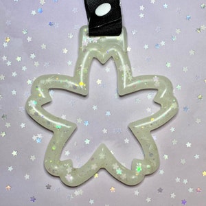 Könnte beinhalten: Ein durchscheinender, blütenförmiger Schlüsselanhänger mit eingebetteten Glitzer und kleinen blütenförmigen Akzenten. Der Schlüsselanhänger ist an einem schwarzen Riemen mit einem weißen Knopfverschluss befestigt. Der Hintergrund ist helllila mit verstreuten sternförmigen Konfetti.