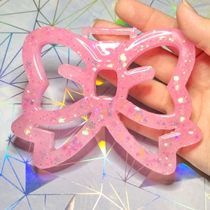 May include: A pink, bow-shaped resin charm with glitter embedded throughout. The charm has a loop at the top for hanging. The bow design features a series of curved lines and open spaces. The charm is held in a hand against a geometric background.