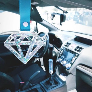 May include: A clear, diamond-shaped car charm with iridescent glitter suspended within. It hangs from a light blue strap attached to the car's interior. The car's dashboard, steering wheel, and gear shift are visible.