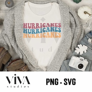 Könnte beinhalten: Ein weißes T-Shirt mit dem Text "HURRICANES", der dreimal in einer Retro-Schrift wiederholt wird. Der Text ist in den Farben Rosa, Blau und Orange gehalten. Das T-Shirt liegt auf einem grauen Cardigan und einer blauen Jeans.
