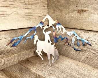 May include: Metal wall art depicting a cowboy riding a bucking bronco in front of a mountain range. The art is made of steel with a blue and purple iridescent finish.