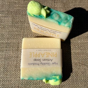 Puede incluir: Dos barras de jabón hechas a mano con un diseño de piña. El jabón es blanco con remolinos verdes y una piña en la parte superior. La etiqueta dice "Hyer Quality Products Artisan Soap PINEAPPLE Hand Made in Ogden, UT".