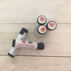 Könnte beinhalten: Vier kleine schwarze Lippenbalsam-Tuben mit Himbeergeschmack. Die Lippenbalsam-Tuben haben ein weißes Etikett mit einem Himbeerdesign und den aufgedruckten Wörtern "Raspberry" und "HYER".