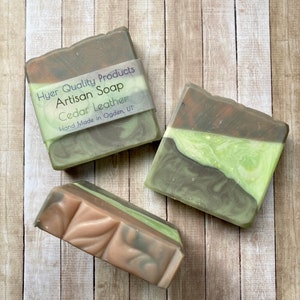 May include: Three bars of artisan soap with a layered design. The soap features shades of brown, green, and tan. One bar is wrapped with a label that reads "Hyer Quality Products Artisan Soap Cedar Leather Hand Made in Ogden, UT."