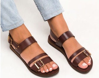 Puede incluir: Sandalias de cuero marrón con dos tiras y un cierre de hebilla. Las sandalias tienen una suela plana y las lleva una persona con pantalones vaqueros.