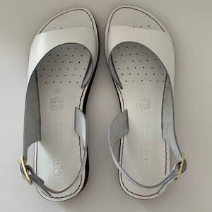 Puede incluir: Un par de sandalias blancas de cuero con cierre de hebilla. Las sandalias tienen un dise&ntilde;o sencillo y est&aacute;n hechas de cuero genuino. Las sandalias est&aacute;n etiquetadas con "CRM SHOES" y "VERO CUOIO".