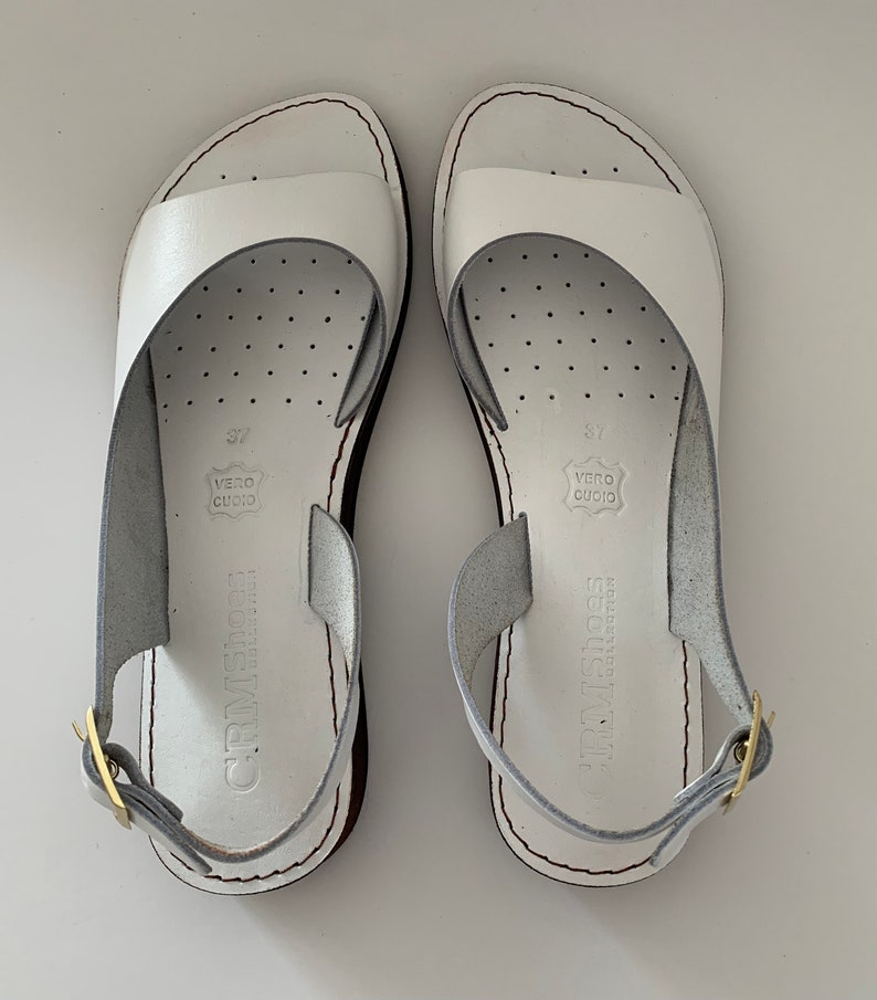 Puede incluir: Un par de sandalias blancas de cuero con cierre de hebilla. Las sandalias tienen un dise&ntilde;o sencillo y est&aacute;n hechas de cuero genuino. Las sandalias est&aacute;n etiquetadas con "CRM SHOES" y "VERO CUOIO".