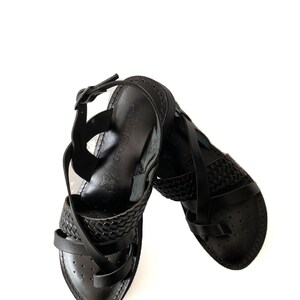 May include: Black leather sandals with a woven strap design. The sandals have a buckle closure and a pointed toe.