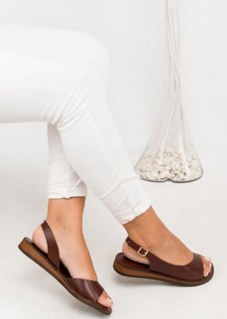 Puede incluir: Sandalias de cuero marr&oacute;n con una correa de hebilla y un dedo del pie abierto. Las sandalias tienen una suela plana y una correa de slingback.