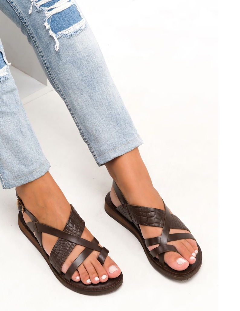 May include: Brown leather sandals with criss-cross straps and a buckle closure. The sandals have a flat sole and a toe loop.