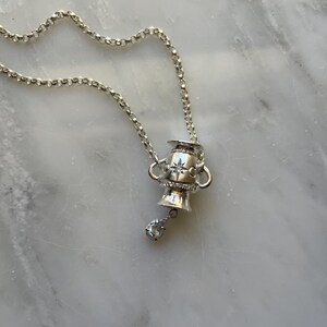 Puede incluir: Un collar de plata con un colgante en forma de copa peque&ntilde;a con un dise&ntilde;o de estrella y un solo cristal transparente que cuelga de la parte inferior.