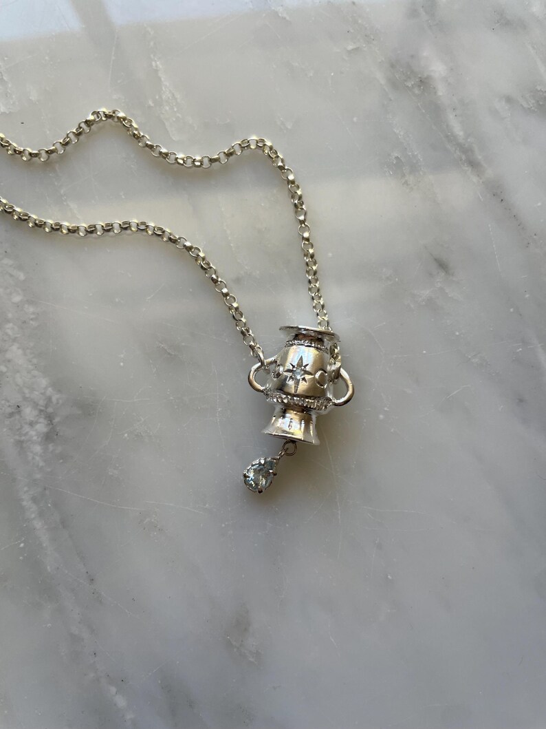 Puede incluir: Un collar de plata con un colgante en forma de copa peque&ntilde;a con un dise&ntilde;o de estrella y un solo cristal transparente que cuelga de la parte inferior.