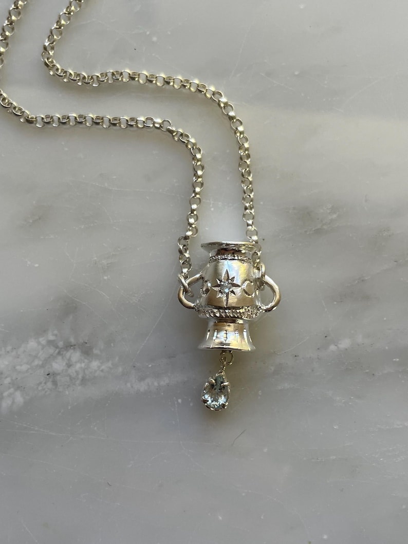 Puede incluir: Un collar de plata con un colgante de urna detallado y una piedra de aguamarina en forma de l&aacute;grima colgante.
