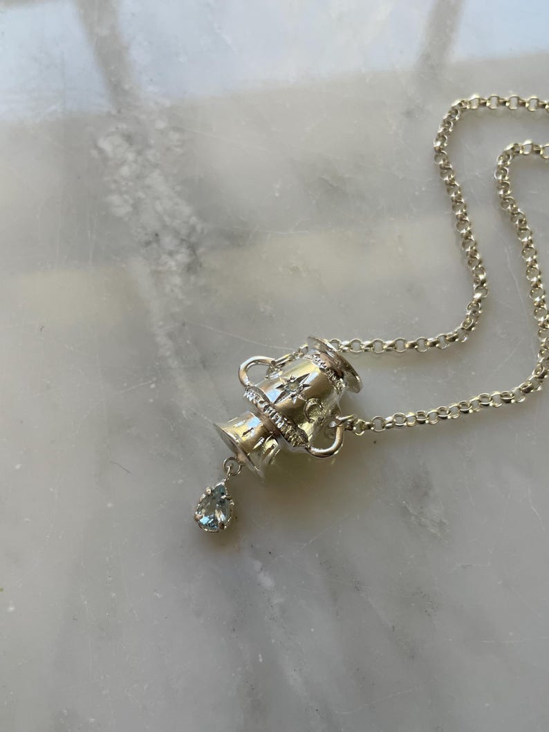 Puede incluir: Un collar de plata con un colgante de urna detallado y una piedra preciosa azul claro en forma de l&aacute;grima.