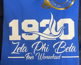 May include: Royal blue sweatshirt with white text. The design includes the number "1920", the words "Zeta Phi Beta", and "Finer Womanhood". A gold emblem is in the upper right corner.