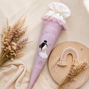 Könnte beinhalten: Eine lavendelfarbene Schultüte mit einer Ballerina-Illustration und dem Namen "Sara". Die Tüte hat einen weißen Aufsatz mit rosa Rüschen und ist mit einer Schnur gebunden. Ein kleiner Makramee-Regenbogen und Trockenblumen sind ebenfalls vorhanden.