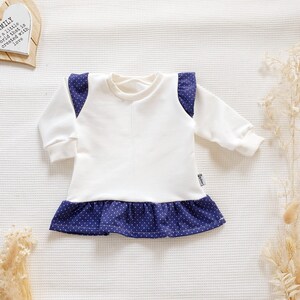 Op de afbeelding: Witte babyjurk met lange mouwen en een blauwe ruche met witte stippen. De jurk heeft blauwe stof met witte stippen op de schouders.