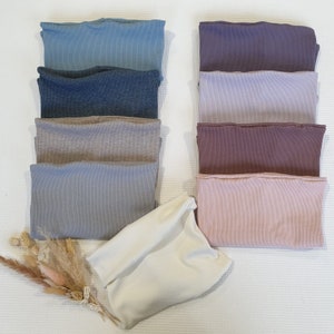 Peut inclure: Plusieurs hauts tubes côtelés pliés, dans des tons de bleu, de gris et de violet. Un haut tube crème est plié en bas, avec des fleurs séchées à gauche. Les hauts sont disposés sur une surface blanche texturée.