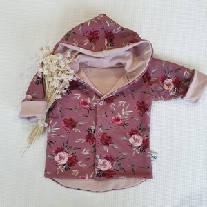 Könnte beinhalten: Eine rosa Kapuzenjacke mit einem Blumendruck mit roten und rosafarbenen Rosen. Die Jacke hat einen Knopfverschluss und ist mit einem weichen, rosa Stoff gefüttert.