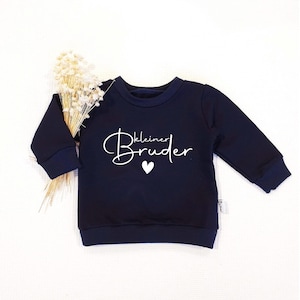 Könnte beinhalten: Marineblaues Baby-Sweatshirt mit dem Schriftzug "kleiner Bruder" in weißer Schrift und einem kleinen weißen Herz. Das Sweatshirt hat marineblaue Bündchen und einen Saum. Links vom Sweatshirt ist ein kleiner Strauß Trockenblumen platziert.