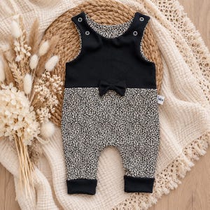 Op de afbeelding: Een mouwloze romper met een effen zwart lijfje, een kleine zwarte strik in de taille en een luipaardprint op het onderste gedeelte en de boorden. Het kledingstuk is voorzien van vier zilverkleurige metalen drukknopen op de schouders.