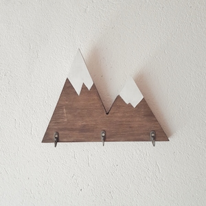 Puede incluir: Porta llaves de madera con forma de montaña con tres ganchos. La montaña está pintada de marrón con picos de nieve blancos.