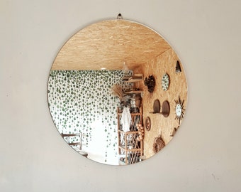 May include: A round mirror with a wooden frame reflects a room with a patterned wallpaper, a wooden shelf with decorative items, and a small table with a chair.