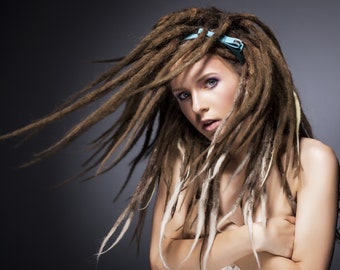 May include: A woman with long, brown dreadlocks and a light blue headband. Some dreadlocks are highlighted with blonde. The woman is looking at the camera with a neutral expression. The background is a dark grey.