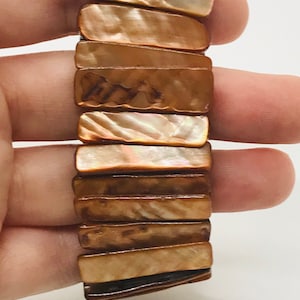 Könnte beinhalten: Ein braunes und weißes Muschel-Armband mit einem geschichteten Design. Das Armband besteht aus rechteckigen Muschelteilen, die übereinander gestapelt sind.