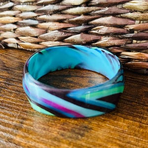 Puede incluir: Una pulsera tipo brazalete ancha con un patrón jaspeado de remolinos en turquesa vibrante, morado intenso y verde brillante. La superficie lisa y pulida del material de resina crea un diseño fluido y continuo alrededor de toda la circunferencia.