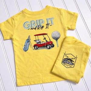 May include: Yellow short-sleeve t-shirt with the text "Grip It and Rip It" and a golf-themed graphic. The graphic includes a golf cart, golf ball, and golf bag. A matching folded shirt with a logo is also present.