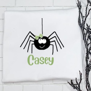 May include: White t-shirt featuring a black spider graphic with green bows. The spider has a smiling face. The name "Casey" is written in green below the spider.