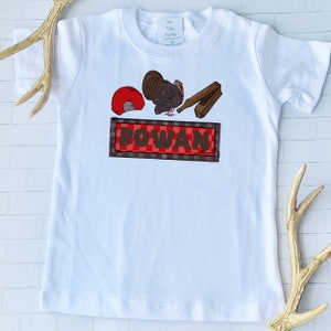 Può includere: Maglietta bianca con un design a tema del Ringraziamento di colore rosso, marrone e nero. Il design presenta un tacchino, un secchio rosso, un bastone di legno e un cartello che dice "ROWAN".