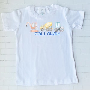 May include: White short-sleeved t-shirt featuring a design of construction vehicles in a watercolour style. The vehicles include a backhoe, cement mixer, and a roller. The name "CALLAWAY" is printed in blue below the vehicles.