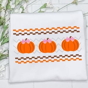 May include: White fabric with a brown and orange zig-zag pattern and three orange pumpkins with pink flowers on top.