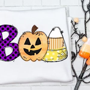 May include: A white t-shirt with a purple polka dot "BOO" design. The "O" is replaced with a cartoon pumpkin with a jack-o-lantern face. The "B" is replaced with a cartoon candy corn with yellow, orange, and white stripes.