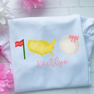 May include: White baby onesie with a yellow watercolor outline of the United States, a red golf flag, a white golf ball with a pink bow, and the name "Kaitlyn" in pink lettering.