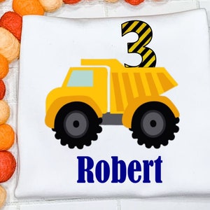 May include: A white t-shirt featuring a yellow dumper truck with the number 3 on top. The truck has black tyres and yellow stripes. The name Robert is printed in blue below the truck.