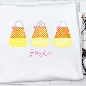 Pode incluir: Camiseta branca com três desenhos de milho doce bordados e o nome "Josie" em letras cor-de-rosa.