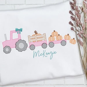 May include: A pink tractor with a teal bow pulls a wagon full of orange pumpkins. The wagon has a wooden slatted bed. The text "Kenzie" is embroidered below the tractor.