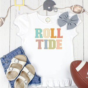 May include: White t-shirt with a colorful "Roll Tide" graphic. The shirt has a ruffled hem and short sleeves. The shirt is laid out on a wooden surface with a football, a football helmet, and a pair of sandals.