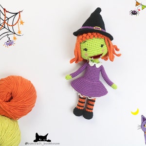 Puede incluir: Una mu&ntilde;eca de bruja de crochet con piel verde, cabello naranja, un vestido morado y medias a rayas negras y naranjas. La mu&ntilde;eca lleva un sombrero de bruja negro con una banda marr&oacute;n. La mu&ntilde;eca est&aacute; de pie sobre un fondo blanco.