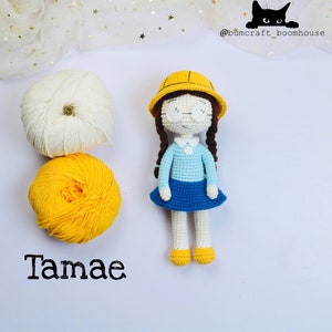 Puede incluir: Una muñeca de crochet con un casco amarillo, una camisa azul y una falda azul. La muñeca tiene el pelo castaño en trenzas y lleva gafas. La muñeca está sentada sobre una superficie blanca con dos ovillos de lana, uno blanco y otro amarillo.