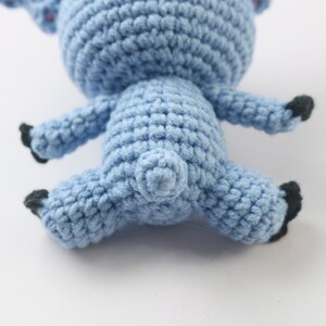 May include: A blue crocheted stuffed animal in the shape of a pig. The pig is lying on its back with its legs in the air. The pig has black eyes and a black nose.