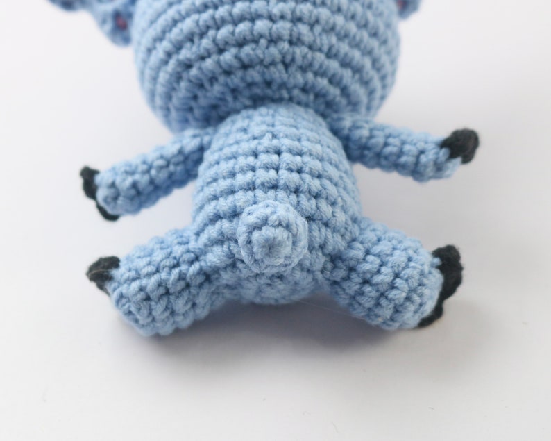 May include: A blue crocheted stuffed animal in the shape of a pig. The pig is lying on its back with its legs in the air. The pig has black eyes and a black nose.