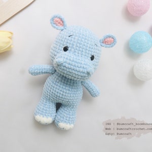 May include: A light blue crocheted hippopotamus toy with pink ears and white feet. The toy is sitting on a white background with some decorative balls.