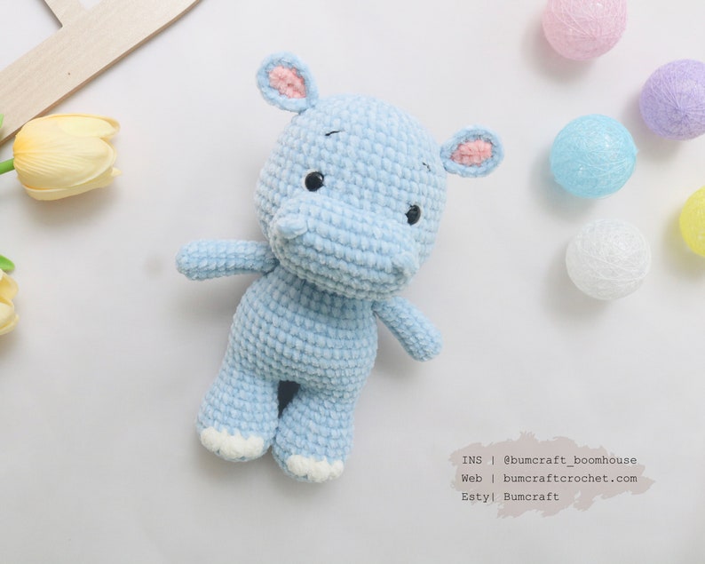 May include: A light blue crocheted hippopotamus toy with pink ears and white feet. The toy is sitting on a white background with some decorative balls.