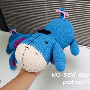 Könnte beinhalten: Ein handgefertigtes blaues Eeyore-Plüschtier aus Häkelarbeit mit weißer Schnauze, schwarzen Details und rosa Akzenten. Das Plüschtier hat eine schwarze Schleife und eine rosa Schleife am Schwanz. Der Text "NO-SEW Eeyore pattern" ist sichtbar.
