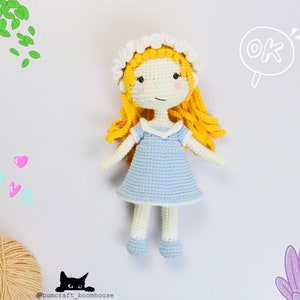 Puede incluir: Una muñeca de crochet con cabello rubio, un gorro blanco de crochet y un vestido azul claro. La muñeca está de pie sobre un fondo blanco con las palabras "OK" y "Love" escritas en el fondo.