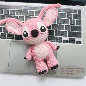 May include: Pink crocheted amigurumi toy with large black eyes and a black nose. The toy is sitting on a laptop keyboard. The text "INS | @bumcraft_boonhouse Web | bumcraftcrochet.com Estyl Bumcraft" is visible on the laptop.